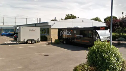 Google A one-storey industrial unit with a grey roof is shown with a black bus and a grey trailer parked on an asphalt car park outside the small building, with bushes and trees to the right of the image and three parked cars in the background on the left.