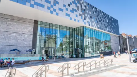 One Plymouth/Art Fund/PA Media A stone, glass-fronted and different-coloured panels run across the box-shaped museum. There is lots of paving outside, steps and hand rails, and a few people hanging around.