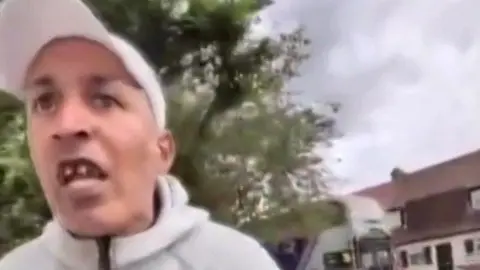 Phone footage of a man in a white cap looking angry, leaning towards the person holding it.