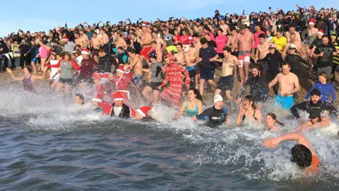 A group of swimmers, some in fancy dress, entering waters off Mersea Island from a beach. The beach is lined with spectators, many taking photos on mobile phones.