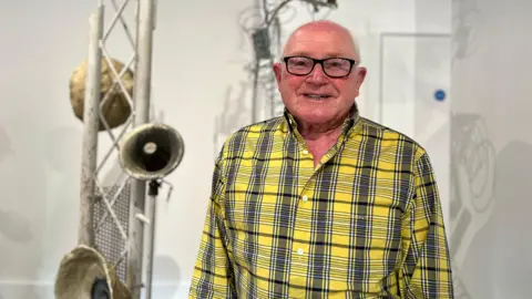 Roy smiles at the camera as he wears a yellow tartan shirt and has glasses. Behind him are old megaphones attached to a building frame