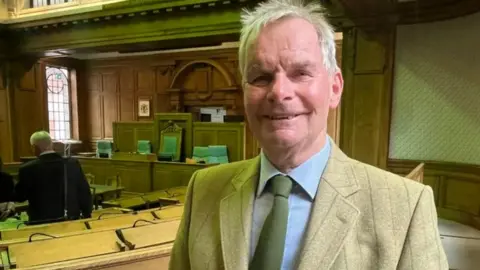 LDRS A head and shoulders view of Martin Hill, the leader of Lincolnshire County Council, who is wearing a beige-coloured blazer, a formal blue shirt and a green tie. He is standing in the council chamber, which has light-wood desks and dark-wood panelling.
