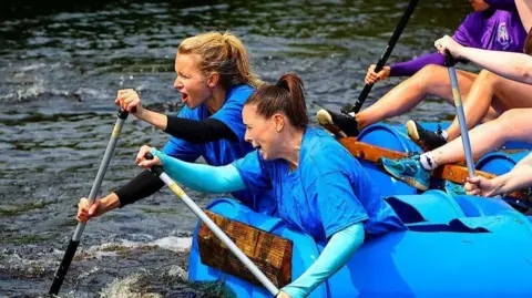 Two woman, one with long blonde hair and one with long brown hair, both in ponytails, are leaning out in front of a boat pulling it along with oars.