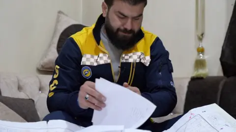 BBC News A man in a White Helmets uniform - dark blue and yellow - looks at documents and maps. 