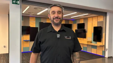 BBC A man with grey hair and a beard is wearing a black short-sleeved shirt. He is looking at the camera and more than half a dozen TV screens mounted on a yellow, green and brown wall behind him are switched off.