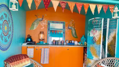 Chrissie Bond The interior of the beach hut has turquoise walls on both sides and the back wall is orange with orange cupboards. Bunting is hanging in the middle and there are two wooden chairs. Artwork has been painted on the walls, including of two peacocks, and there are various tea towels, a kettle, a mirror and pots neatly lined up.