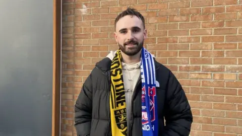 A young white man with dark hair and a bears smiles at the camera wearing a Maidstone United and Ipswich Town scarf and black coat