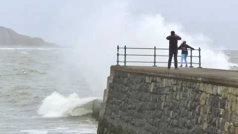 BBC Weather Watcher/snaphappydave Two people watch the stormy seas at the end of a pier in Criccieth, Wales