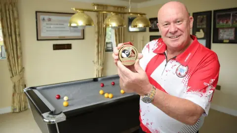 PA Media Neil Jones, 59, from Stoke-on-Trent, with his bronze medal he won as part of the England B1 team which he captained at the European Blackball Championships in Malta in November. Mr Jones won £2.4 million on the National Lottery in 2010 and a pool table was one of his first purchases, going on to be selected for England after honing his skills over the Covid lockdown. Issue date: Monday November 20, 2023. PA Photo. See PA story LOTTERY Pool. Photo credit should read: National Lottery/PA Wire