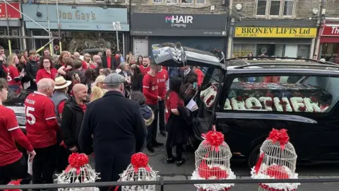 Fiona Lamdin/BBC Mourners dressed in red gather around a hearse ahead of the funeral of Mason Rist in Bristol