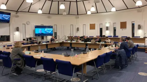 West Northamptonshire councillors - about 11 of them - are sitting around tables in a modern council chamber. A man's face is visible on a television monitor, who is joining the meeting remotely