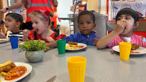 Four nursery pupils sat at a canteen table enjoying their Friday fish and chips dinner at St Mary's Primary in Stoke-on-Trent, including spaghetti hoops. They are in fancy dress for 'aspirations day'.