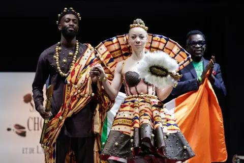 Stephane de Sakutin / AFP Ivorian designer Roger Bango (R) acknowledges the applause after France's Paris 2024 Paralympic flag bearer Nantenin Keita (2nd R) presenting a model created by him during the chocolate dress parade of the opening day of the Salon du Chocolat de Paris 2024 (2024 Paris Chocolate Fair) in Paris.