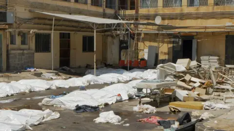BBC / Jon Donnison White plastic sheets cover bodies in the carpark of a hospital in Suweida. There is debris of chairs and hospital beds scattered across the ground too