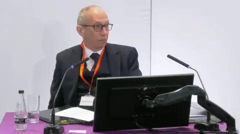 Sir Rob Behrens appearing at the Lampard Inquiry. He sits at a purple-covered desk with two microphones pointing towards him and a computer monitor in front of him. He has a bottle of water and a glass to his right hand side. He is wearing a dark suit with dark waistcoat, dark tie and white shirt. he has dark-rimmed glasses.