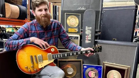 PA Media Auctioneer Luke Hobbs hllding Ian Bairnson's guitar and kneeling near the rest of the equipment