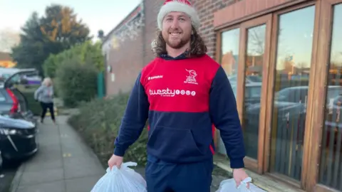 Simon Thake Connor Davidson has long curly hair and is wearing dark blue and red sports clothes and a Santa hat. He is smiling as he lifts two white shopping bags.