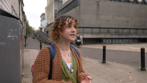 A woman wearing a brown knitted cardigan and a green top. She has brown curly hair and is wearing three necklaces. She is wearing a blue backpack and is looking to the right of the camera.