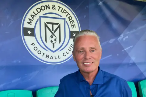 Warren Muggleton/BBC Reality star Barrie Drewitt-Barlow stands next to the spectator seats at Maldon and Tiptree FC.