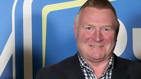 Jim Gracey looking at the camera and smiling. He has short hair and is wearing a black and white checked shirt and a dark blazer