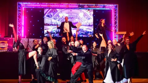 Knot Events/Mark Scott More than a dozen people in smart clothing are stood in front of a stage, with a number of them throwing their arms in the air. There is a brightly lit frame on the stage, with a man and woman looking out towards the audience
