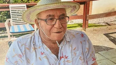 Donald Burgess wears a straw sun hat tied under his chin and a white shirt with yellow and red petal-type shapes on it. He is sitting outside on a patio and is wearing glasses.