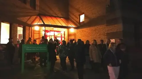 James Avrill/LDRS Residents queuing at night outside brick building with red rails