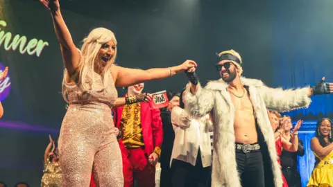 Nick Allan A woman in a sequin catsuit and blonde wig on a stage with one arm in the air and the other holding the hand of a man in a fur coat, dark glasses, silver trainers and leather headband and gloves. The woman is smiling broadly at an unseen audience, the man is looking at her, and other performers around them applaud.