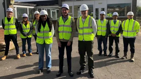 Nine men and women stood in green reflective jackets and hard hats in front of a building with large windows and white panes.