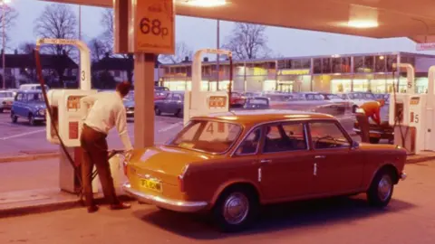 A man filling a car at a petrol station in 1975. He has his back to the viewer, is facing a white petrol pump and is holding the petrol feed which is filling a an orange car. He is wearing a white shirt tucked into brown trousers. Above his car is a sign saying petrol 68p