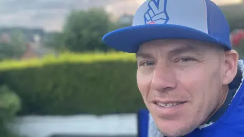 Mark is wearing a blue top and blue baseball cap. He is smiling at the camera. There is a hedge behind in.