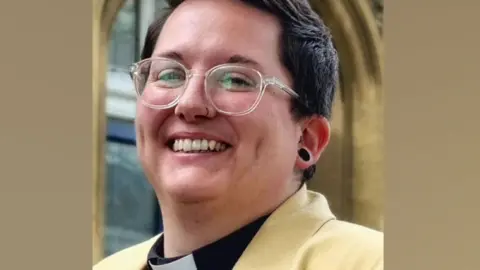Peterborough Cathedral Lex Bradley-Stow who is smiling at the camera with her head slightly tilted back. She has short dark hair and is wearing clear framed round glasses, a yellow jacket and a black shirt with a white dog collar.