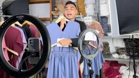 Evo Sya Streamer, Evo Sya, holding a blue summer dress while filming his live stream