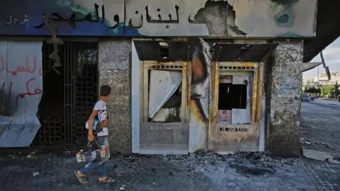 AFP A youth walks past a burnt-down branch of a Lebanese bank in al-Nour Square in Tripoli, Lebanon (12 June 2020)