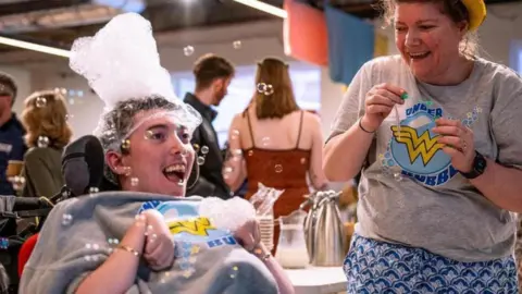 A man sits in a wheelchair. He wears a grey sweatshirt which has the words 'wonder bubbles' on it, in yellow writing. The man has a collection of small bubbles on his head. A woman stands to his left, she wears the same top, but with a blue skirt. She is holding a bubble blower.