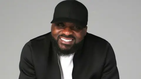 BBC DJ Ace smiling at the camera, he is wearing a black jacket, white t-shirt and black baseball hat.