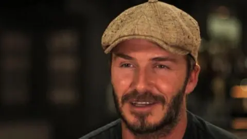 David Beckham wearing a sandy coloured flat cap and dark T-shirt.