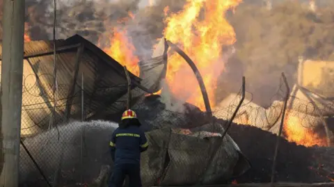 Feuerwehrleute der Environmental Protection Agency (EPA) sprühen Wasser auf ein Gebäude in Penteli, nordöstlich von Athen. Das Gebäude hat ein Wellblechdach und ist größtenteils eingestürzt. 