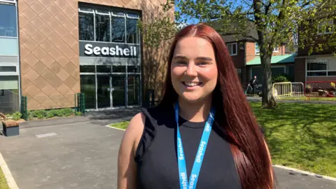Aine Hickey is a teacher at Royal College Manchester. The image is taken outside the Ged Mason building at the Seashell Trust. She is smiling, has long, straight brown hair and is wearing a black sleeveless dress. She has a blue lanyard around her neck.