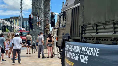 A girl straddles the climbing wall, with two boys either side of her. A small crowd of people have gathered round to watch, and an army vehicle is in the foreground. A banner on its side says: "Join the army reserve in your spare time - recruiting now".