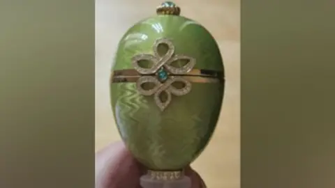 Close-up of a green Fabergé egg ornament featuring a gold band around the centre and a decorative diamond‑patterned design with a small blue‑green gemstone.