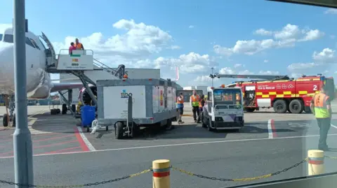 Paul Bartlett Seen through the window, the white plane sits on the tarmac with several vehicles nearby including a fire engine. The side of the plane is open with the steps leading down and some people in high vis vest
