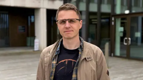 Dr Michael has closely cropped hair and is wearing glasses dressed in a beige jacket over a black graphic T-shirt and a checked shirt, he is standing outside a modern building with glass doors and concrete columns. 