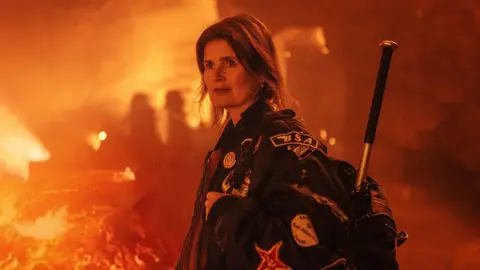 Sophie Aldred in Doctor Who in 2022, dressed in her jacket, stood inside a film set made to look like a volcano, near a stream of lava.