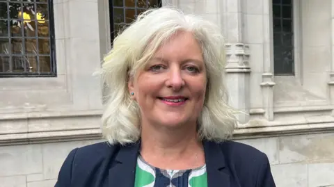 South Devon MP Caroline Voaden smiles at the camera while stood in front of a grey stone building. She has wavy white hair. She is wearing a navy blue blazer and a green, blue and white dress underneath.