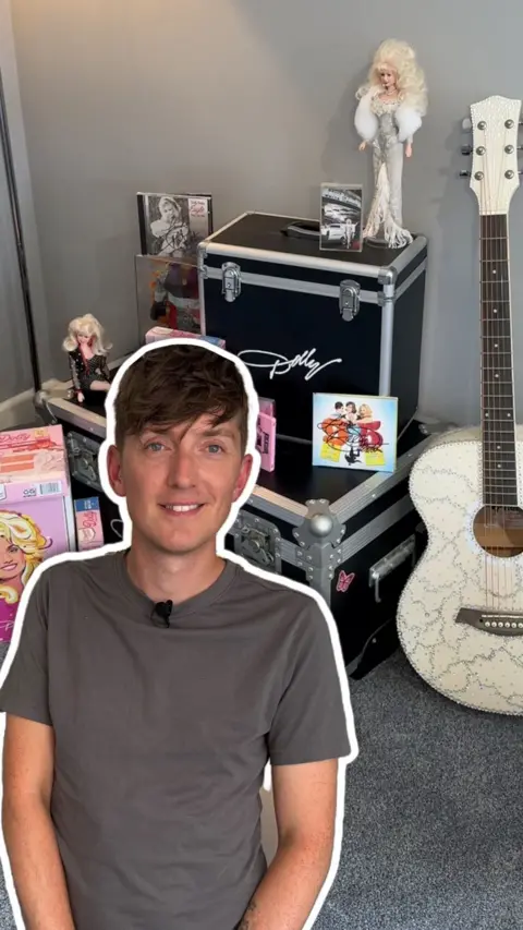 A man with brown hair wearing a grey shirt outlined in white in front of Dolly Parton memorabilia