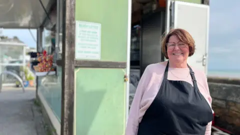 Rhona Hewit, a woman in a light pink sweater and black apron, stands outside a small green food kiosk with an open door. A sign on the wall warns against beach littering and encourages proper bin use to keep Jersey clean. The surrounding area includes some plants and an outdoor setting.