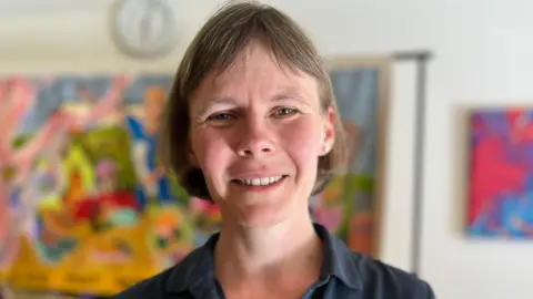 Head and shoulders shot of a smiling woman, wearing a dark blouse. A multi-coloured artwork is hanging on a wall behind her but the background is blurred.
