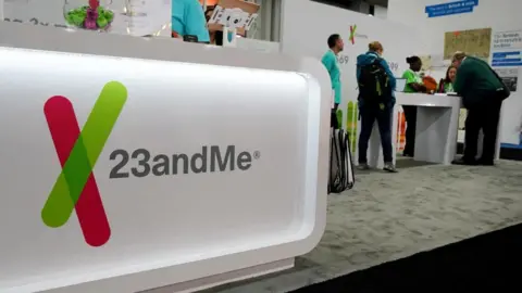 The 23andMe logo is seen at a genealogical convention. The logo is on a bright, white display stand at the front of a carpeted exhibition stall for the company, with people in light blue uniform shirts and others in plain clothes around a table in the background.