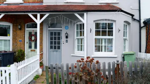 PA Media A suburban terraced house with a bay window and a small front garden surrounded by a weathered and white fence. The house and front door are painted grey. 